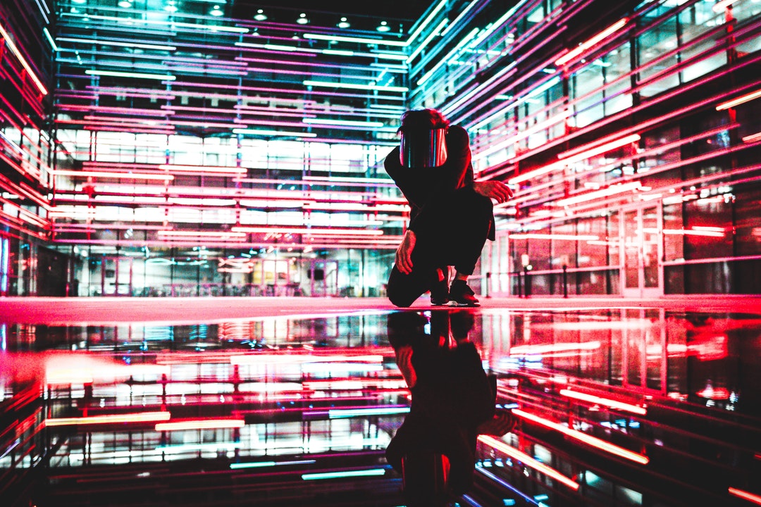 A person in a glossy mask crouching on the reflective floor in a neon-soaked building