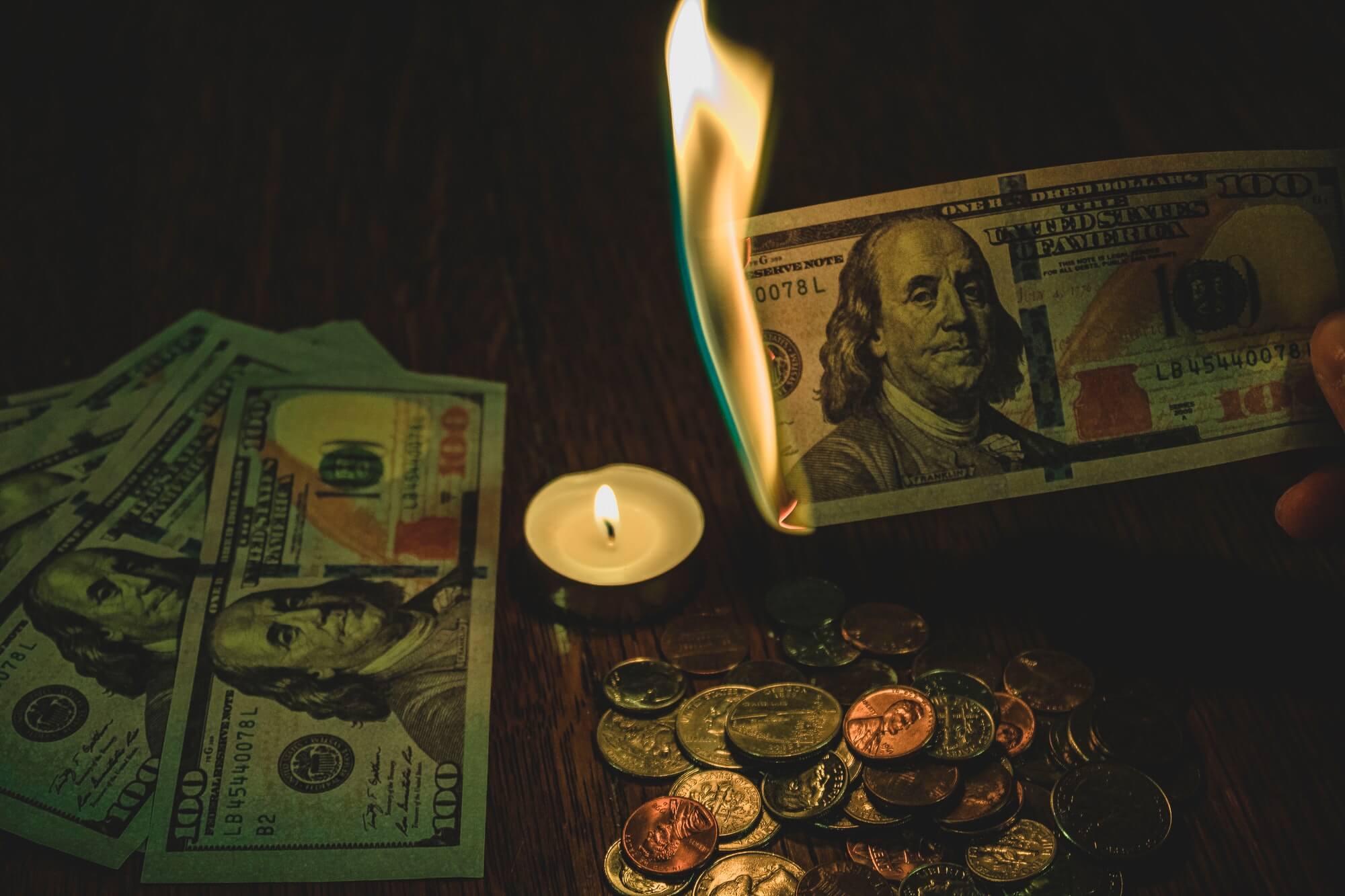 100 dollar bill in flames over candle surrounded by coins and 100 dollar bills. 